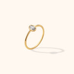 Gold-colored ring with a slim polished band and an oval, clear faceted center in a smooth bezel, tilted against a cream background.