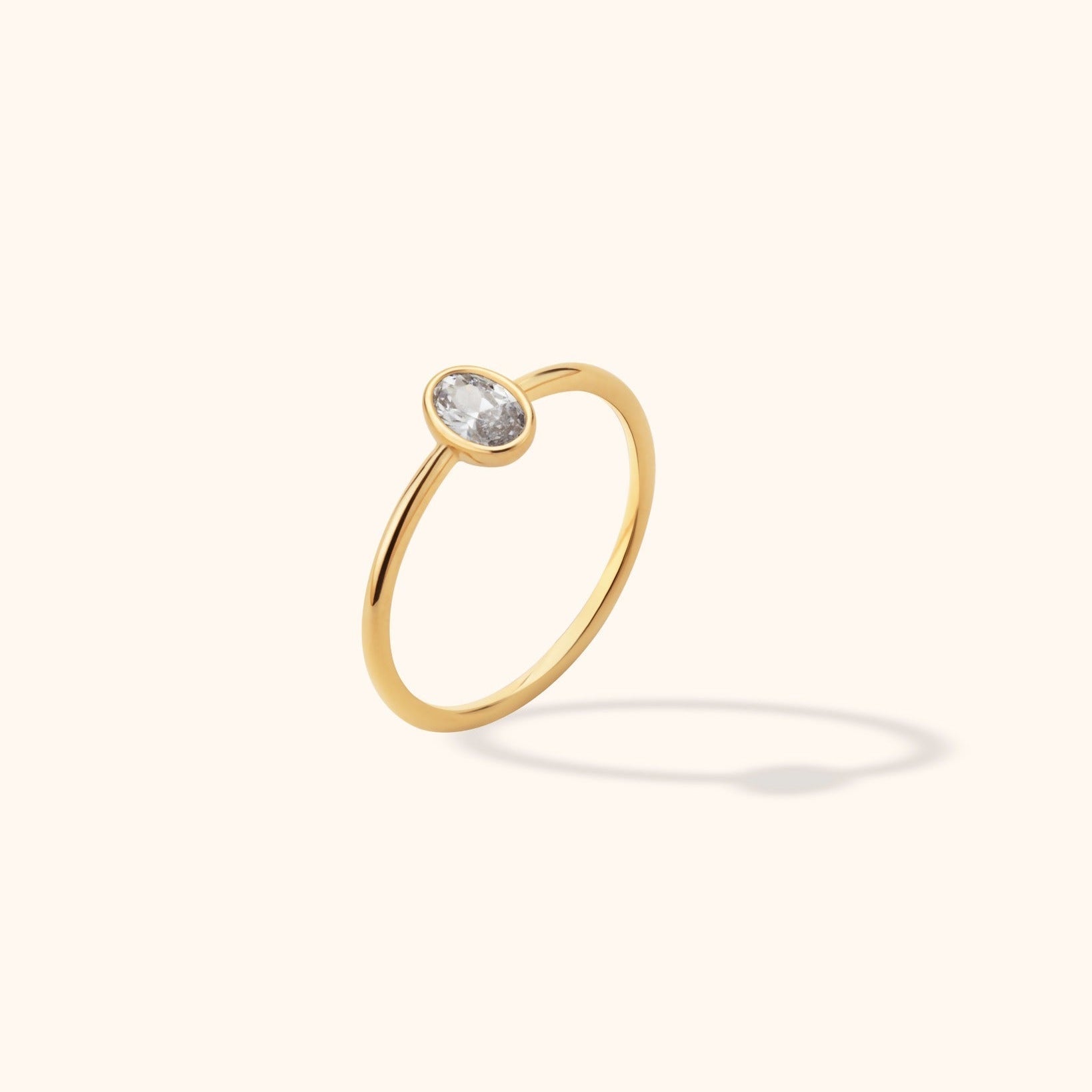 Gold-colored ring with a slim polished band and an oval, clear faceted center in a smooth bezel, tilted against a cream background.