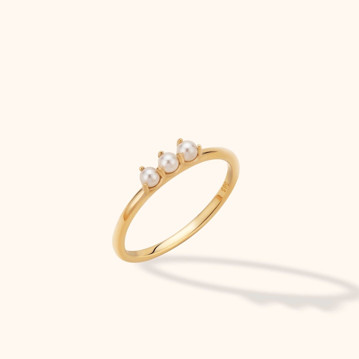 Slim gold-toned ring with three round, white pearl-like beads in prong settings, shown at an angle on a cream background.