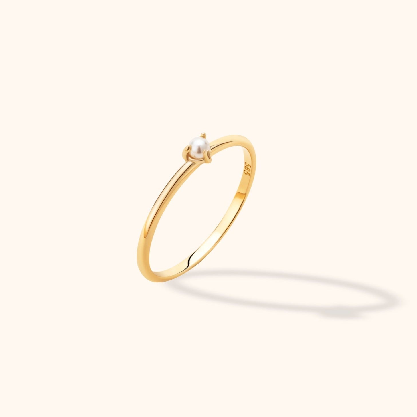 Slim gold-toned ring with a single round white pearl-like bead in a prong setting, tilted against a cream background.