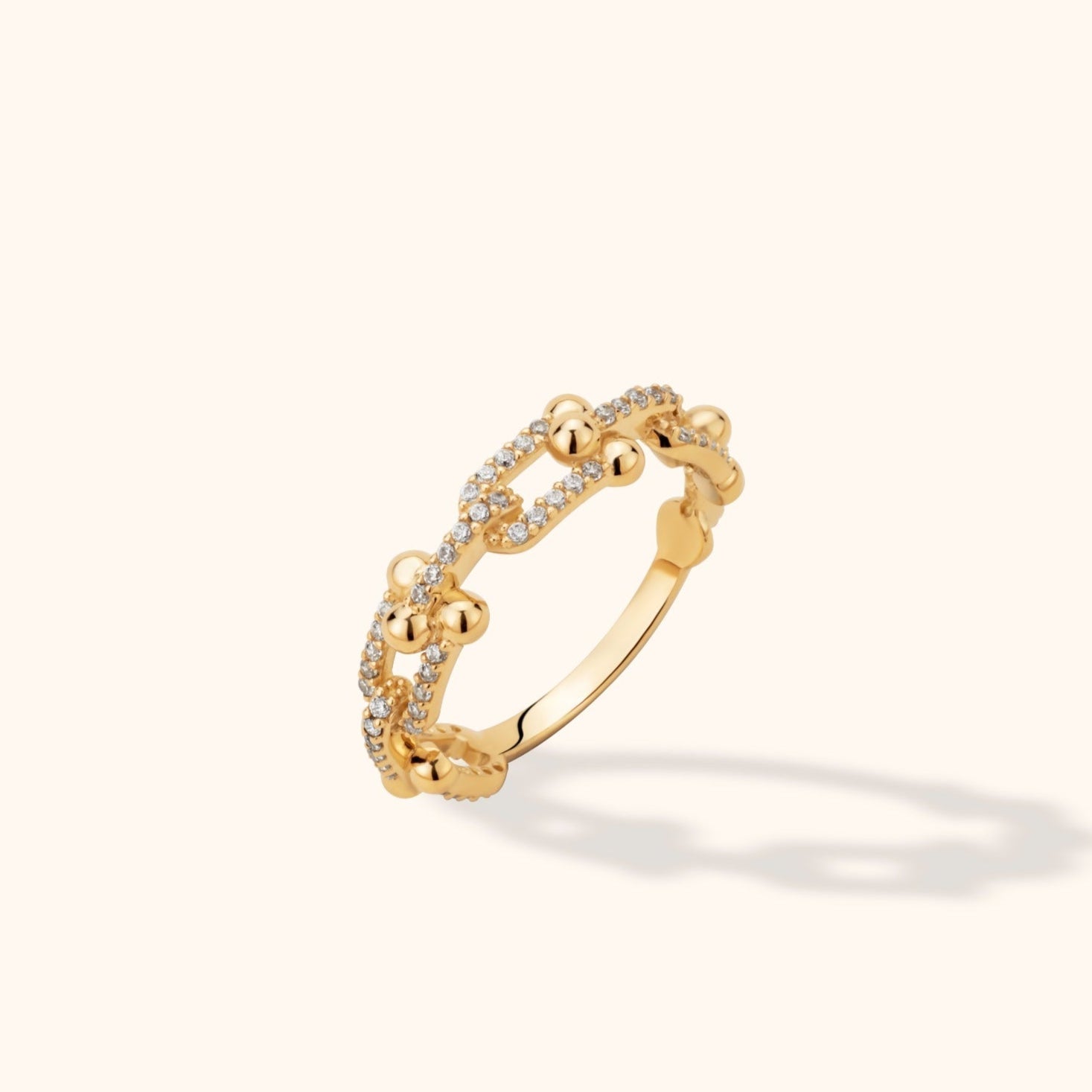 Gold-tone ring with open chain-link shapes, rows of small clear stones and rounded bead details, angled against a cream background.
