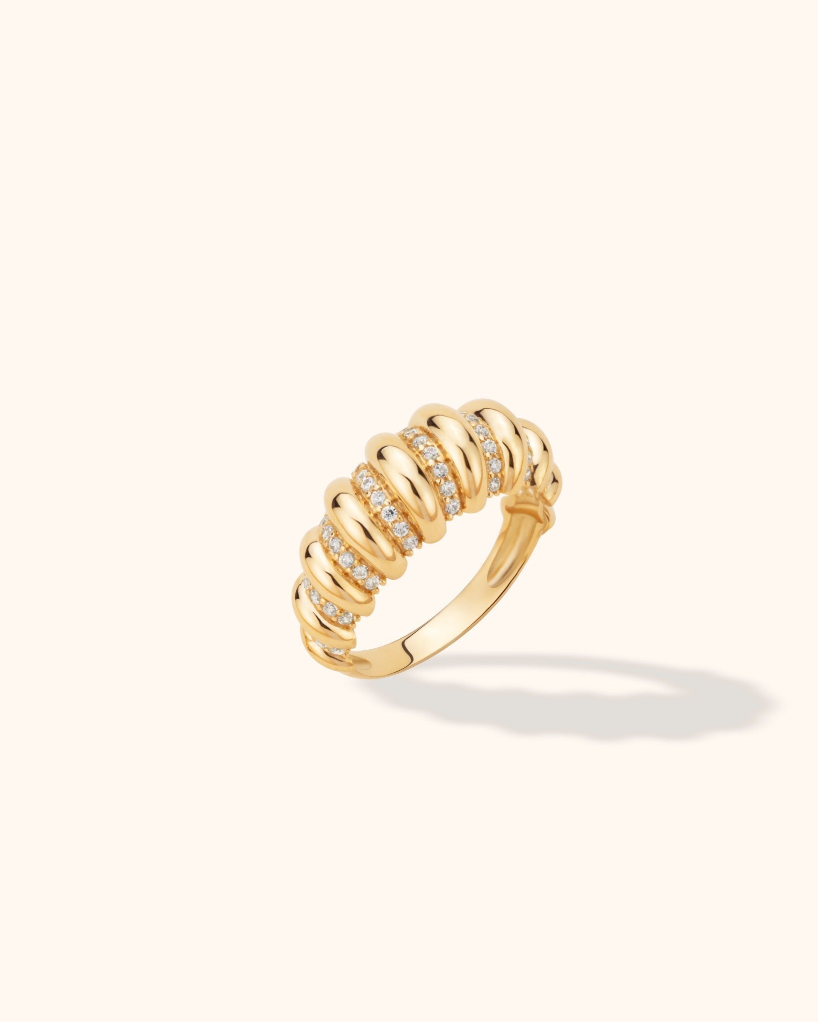 Gold-toned ring with rounded, polished ribs alternating with rows of small clear stones, shown at an angle on a cream background.