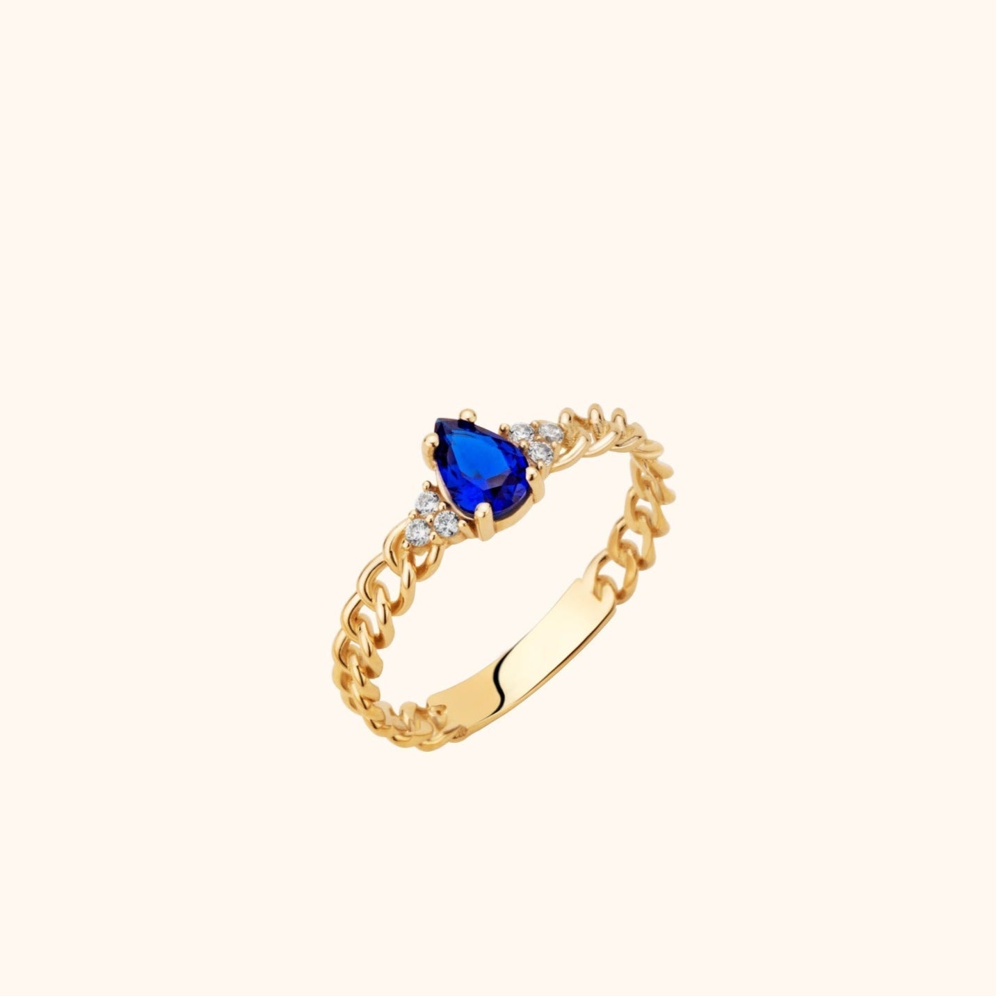Gold-toned chain-link ring with a pear-shaped blue center stone and small clear stones on either side, shown at an angle.