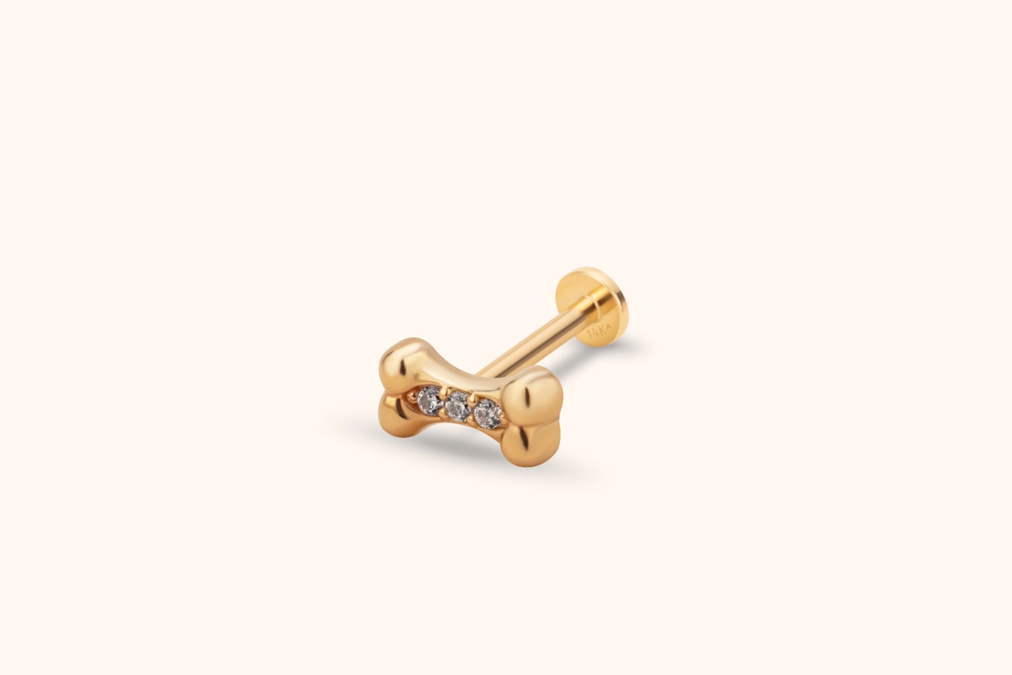 Gold-colored dog bone stud with three round clear stones, a straight post and flat circular back on a cream background.