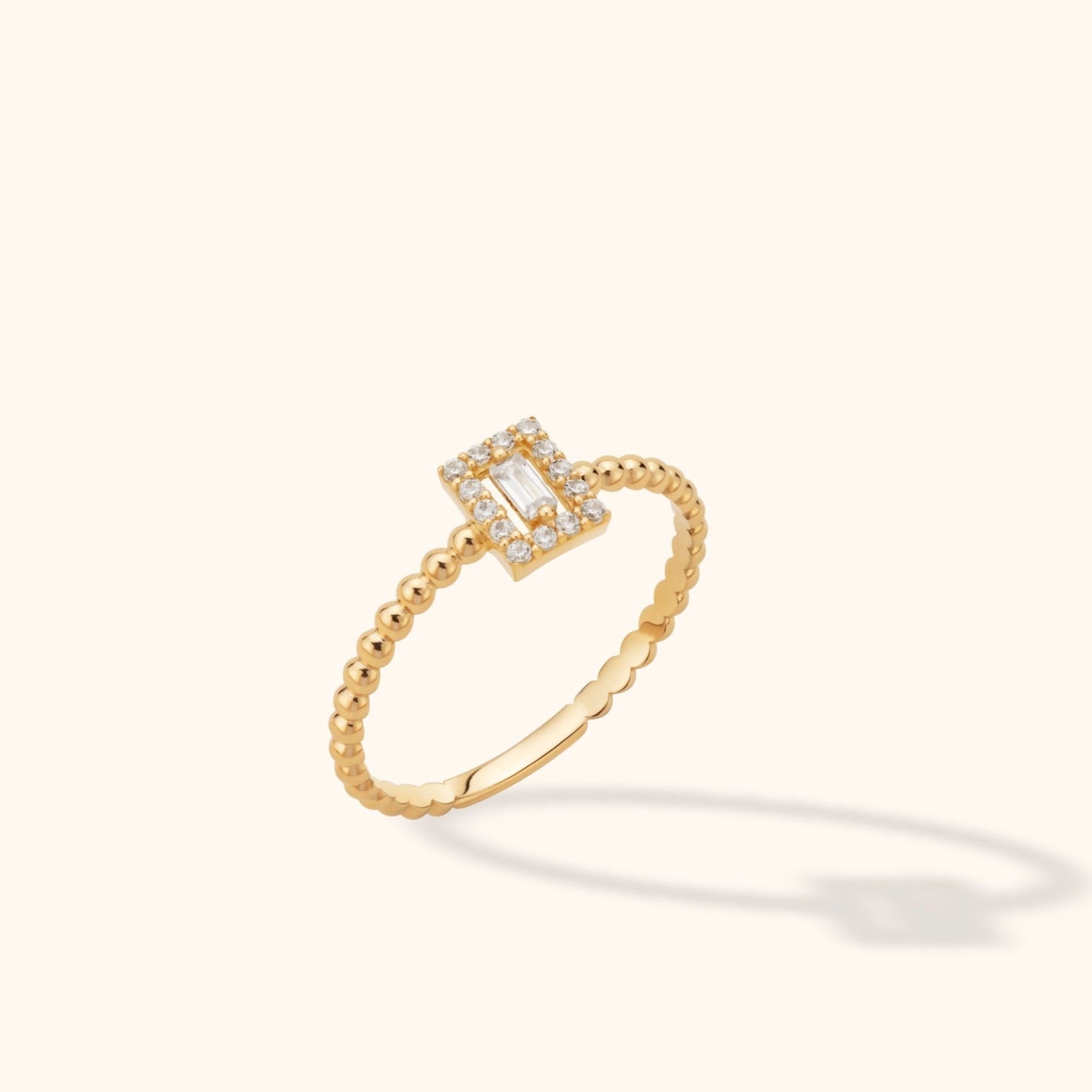 Gold-tone ring with a slim beaded band and a rectangular clear center stone surrounded by a halo of small round clear stones.