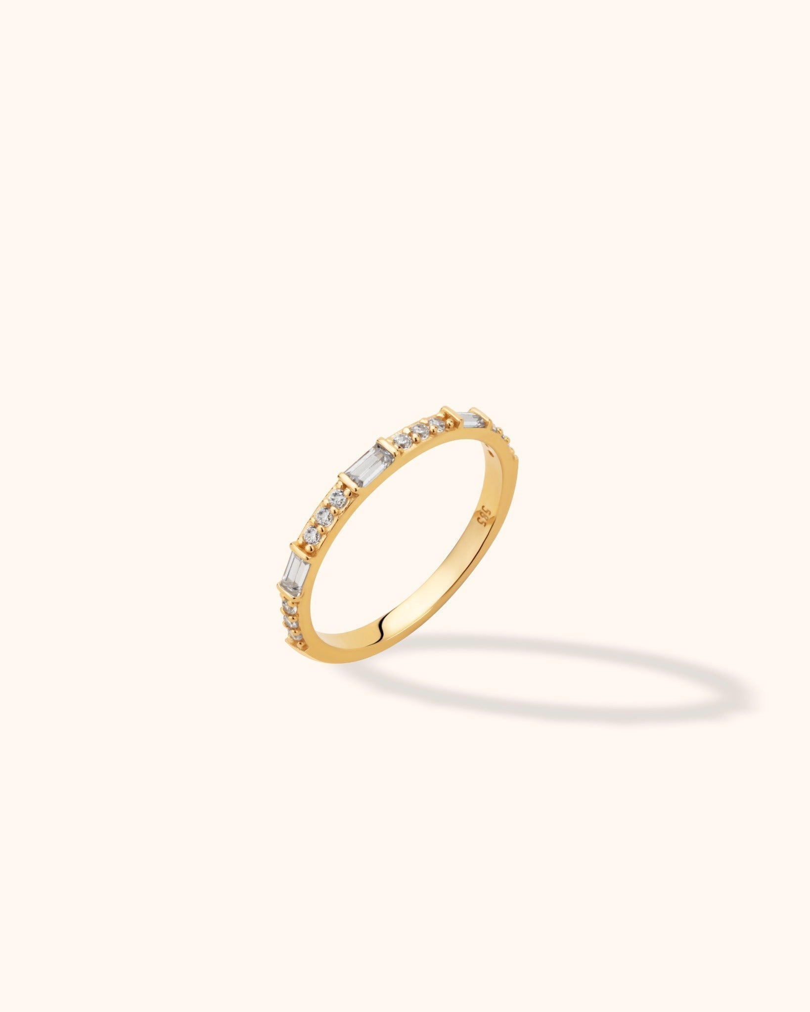 Slim gold-toned ring with clear rectangular stones alternating with groups of round stones, shown at an angle on a cream background.