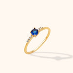 Slim gold-toned ring with a round blue center stone in a prong setting and three small clear stones on each side.