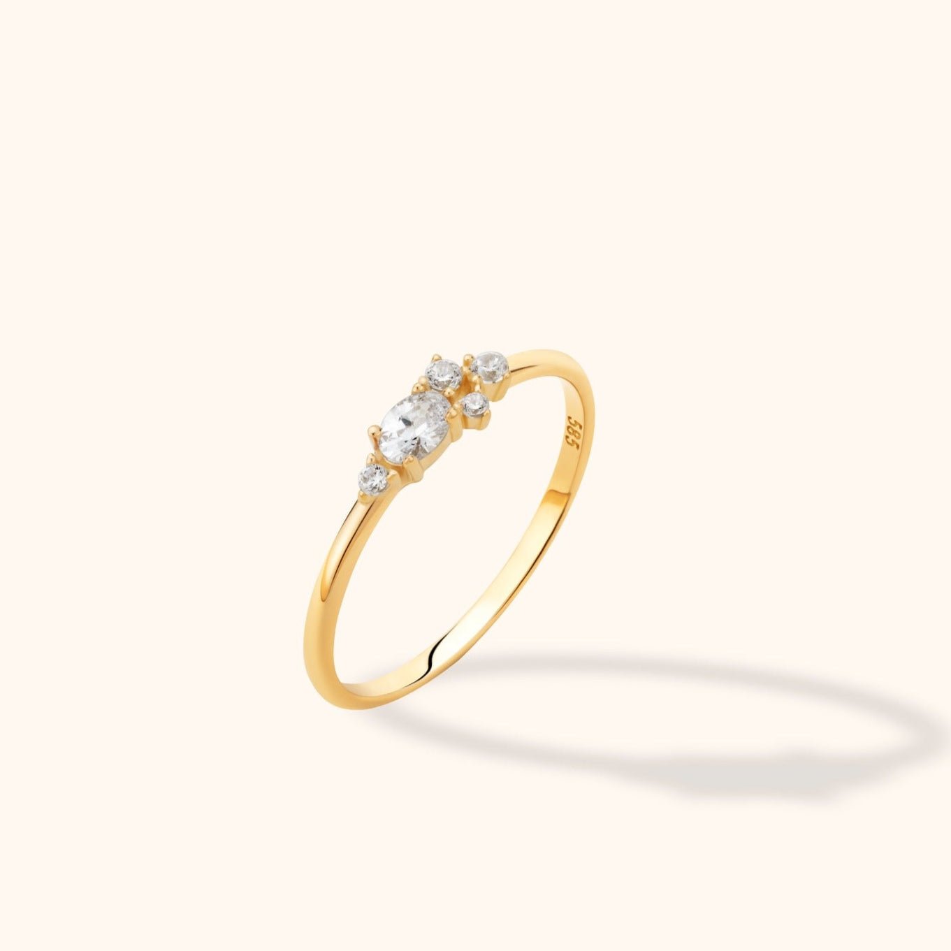 Slim gold-colored ring with an oval clear stone and four smaller round clear stones, shown at an angle on a pale background.