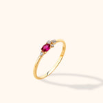 Slim gold-toned ring with an oval deep pink stone and four small clear stones, shown at an angle on a cream background.