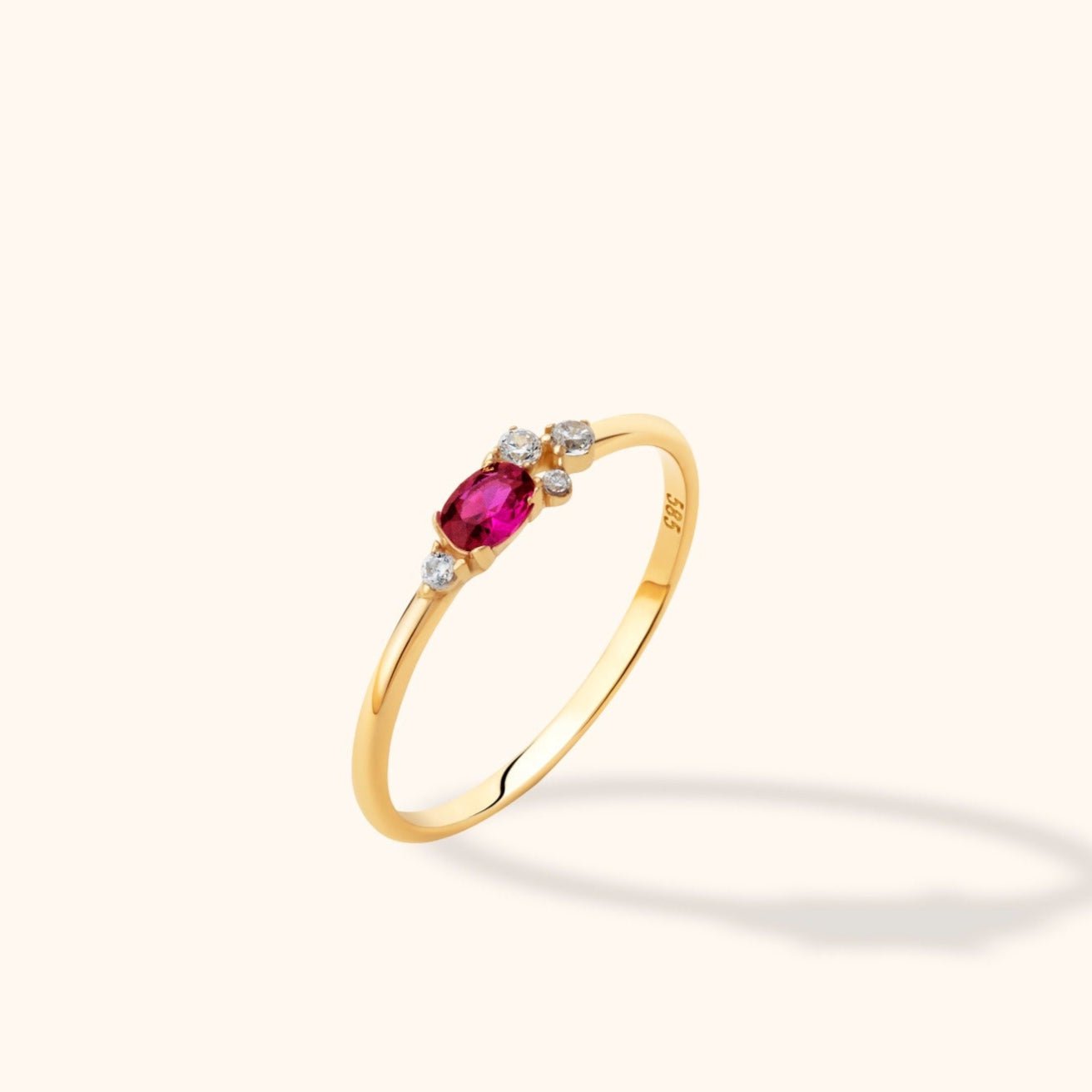 Slim gold-toned ring with an oval deep pink stone and four small clear stones, shown at an angle on a cream background.
