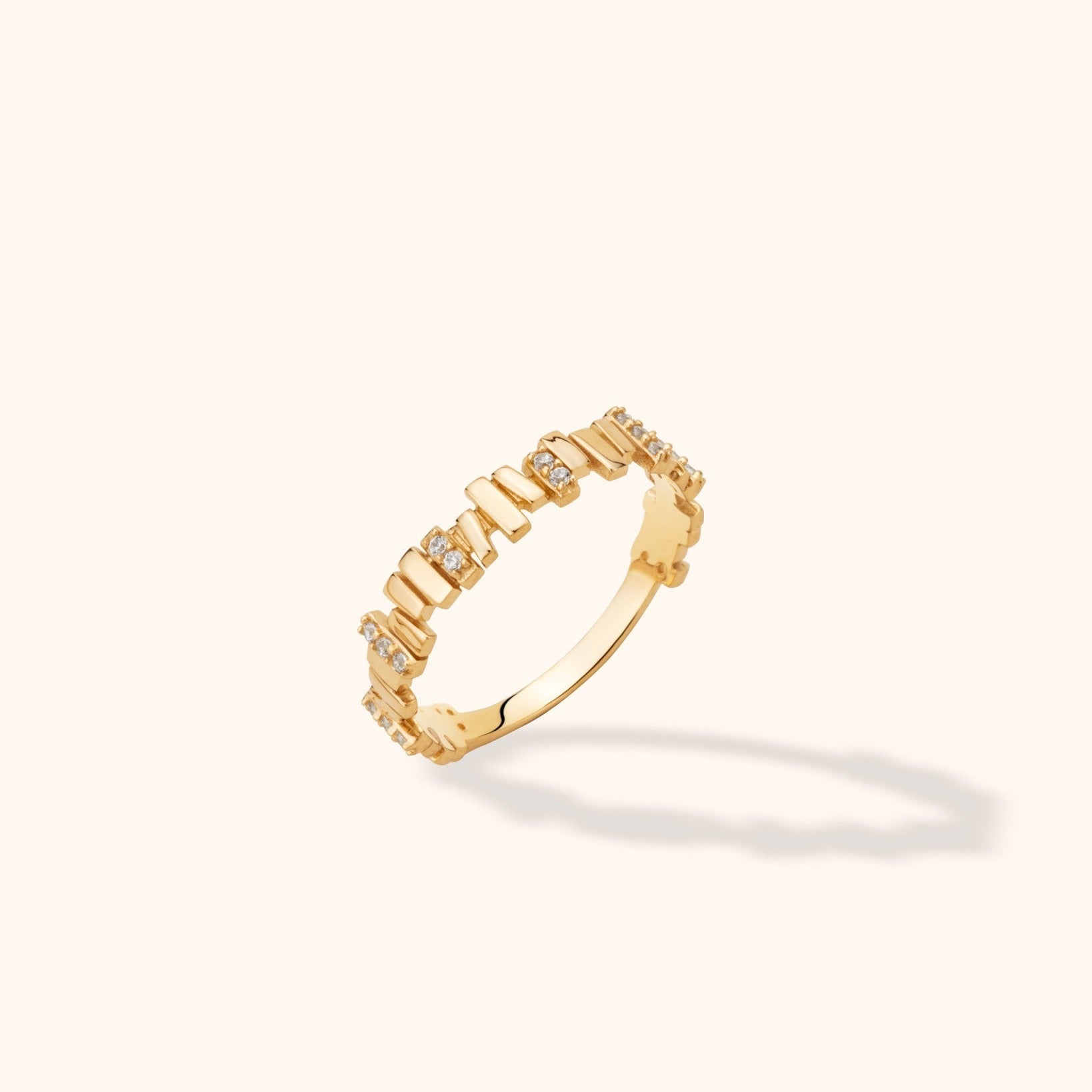 Gold-colored ring with polished geometric segments and small clear stones, shown tilted against a pale cream background.