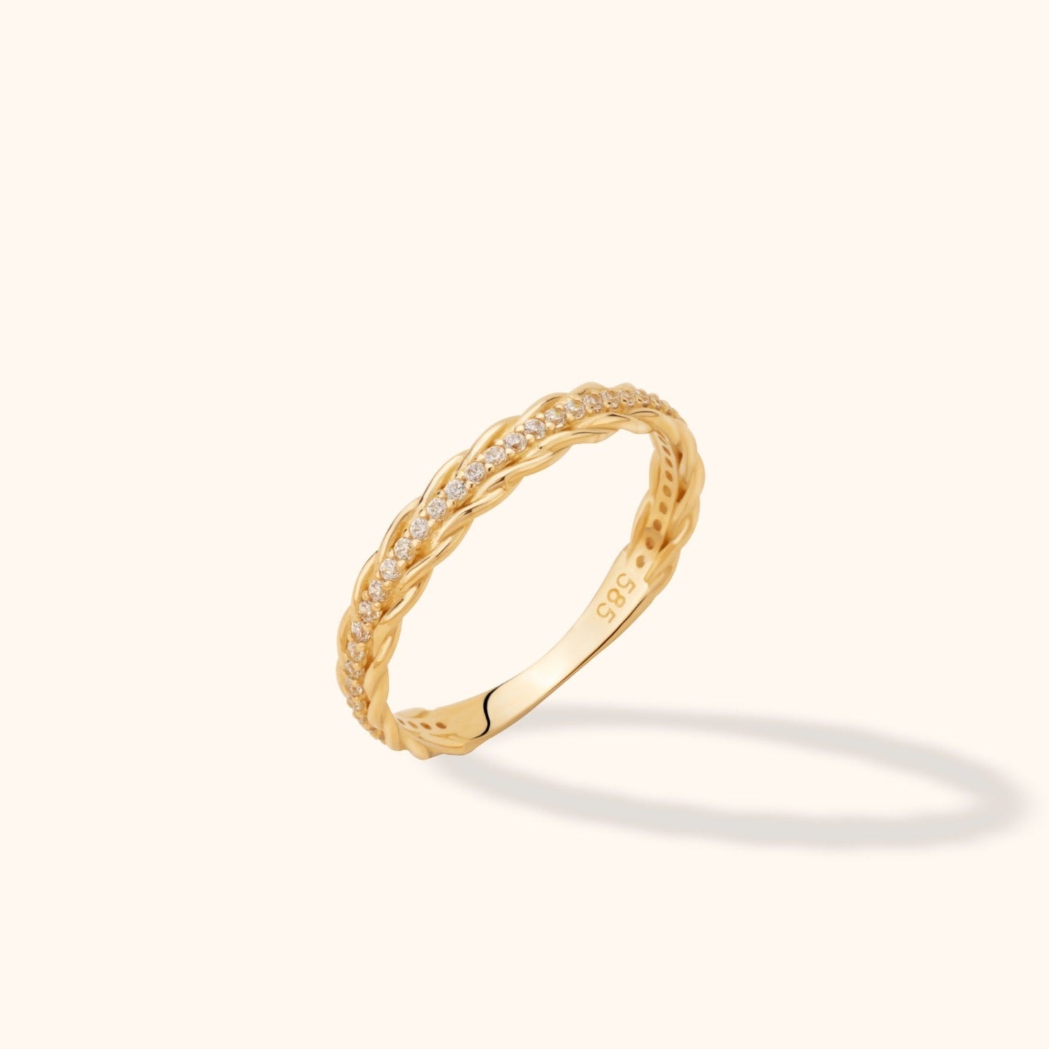 Gold-colored ring with braided edges and a row of small clear stones, shown at an angle on a pale cream background.