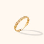 Gold-colored ring with braided edges and a row of small clear stones, shown at an angle on a pale cream background.