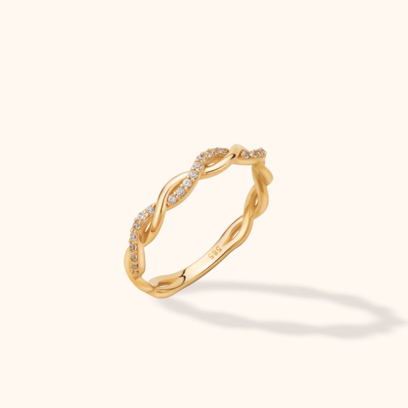 Gold-colored ring with intertwined bands and rows of small clear stones, shown at an angle on a pale cream background.