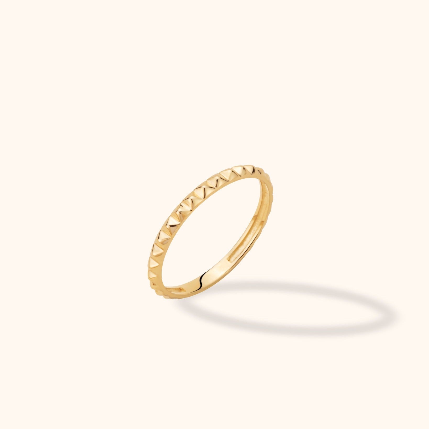 Slim gold-colored ring with raised pyramid-shaped details, shown tilted against a cream background with a soft shadow.