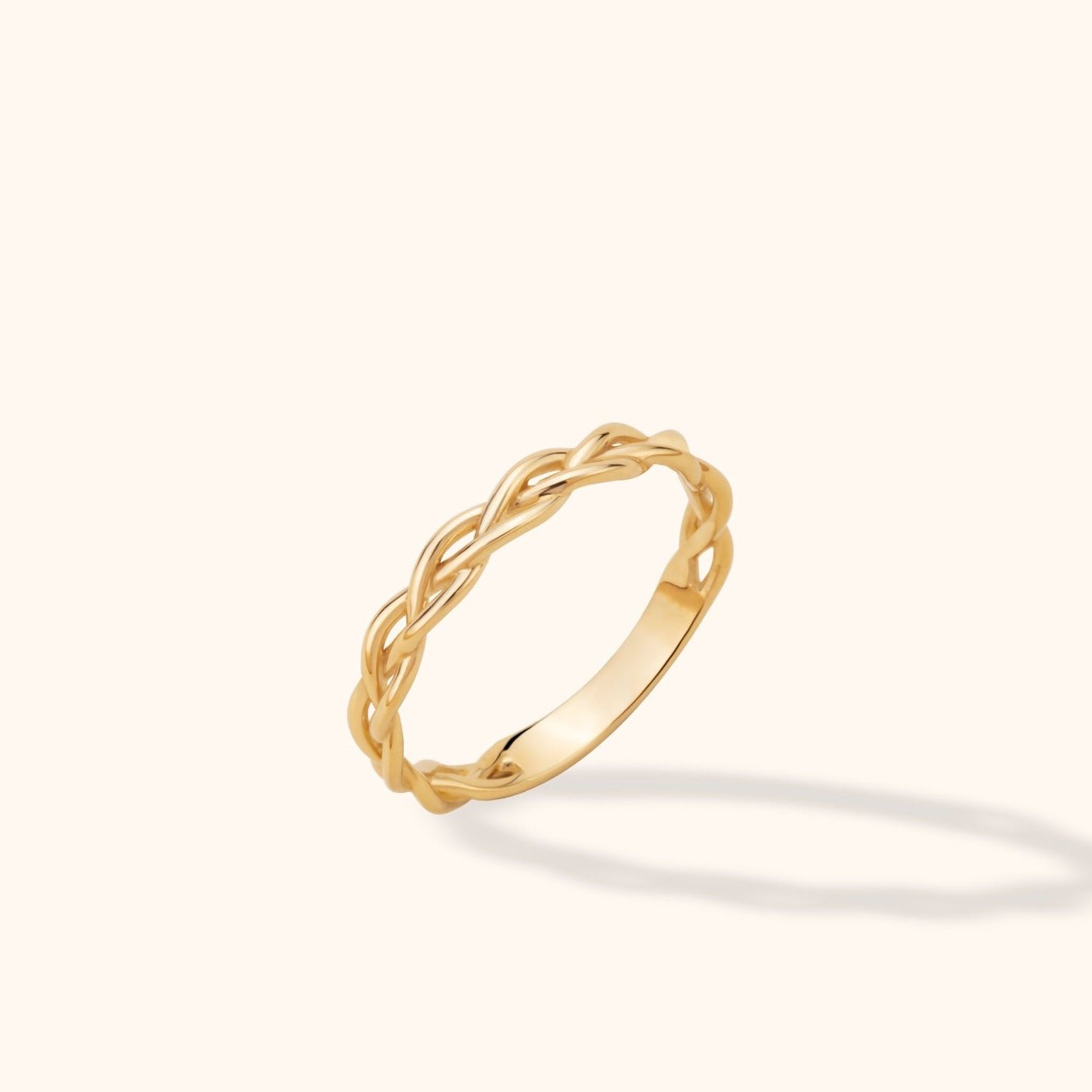 Gold-toned ring with an open braided design and a smooth lower band, tilted against a pale cream background.