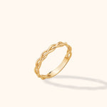 Gold-toned ring with an open braided design and a smooth lower band, tilted against a pale cream background.