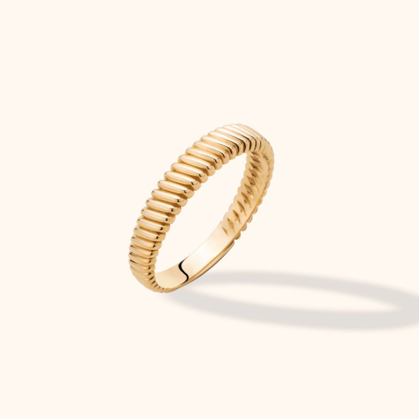 Gold-colored ring with closely spaced rounded ribs, shown tilted on a cream background with a soft shadow.
