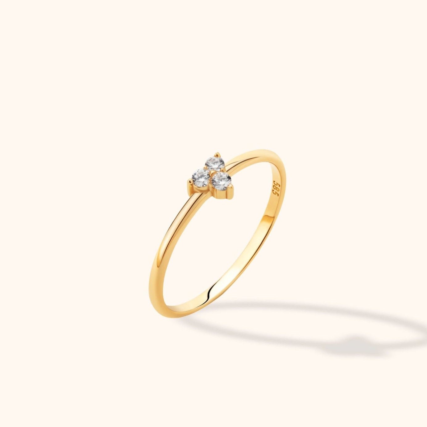 Slim gold-colored ring with three round clear stones in a triangular cluster, shown at an angle on a cream background.