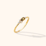 Slim gold-toned ring with an oval olive-brown stone and four small clear accent stones, shown at an angle on a cream background.
