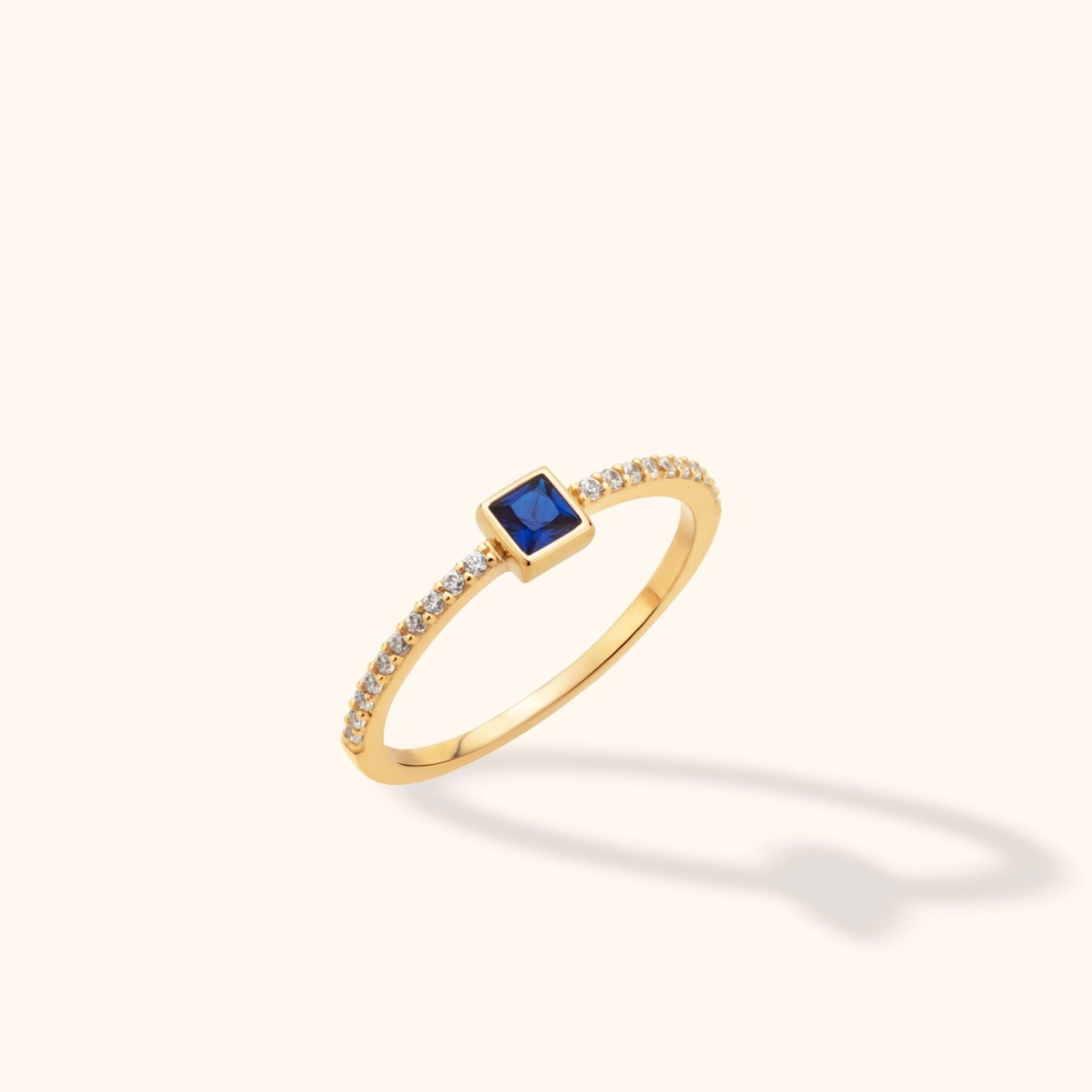 Slim gold-tone ring with a square blue center stone and small clear stones lining the shoulders, shown at an angle.