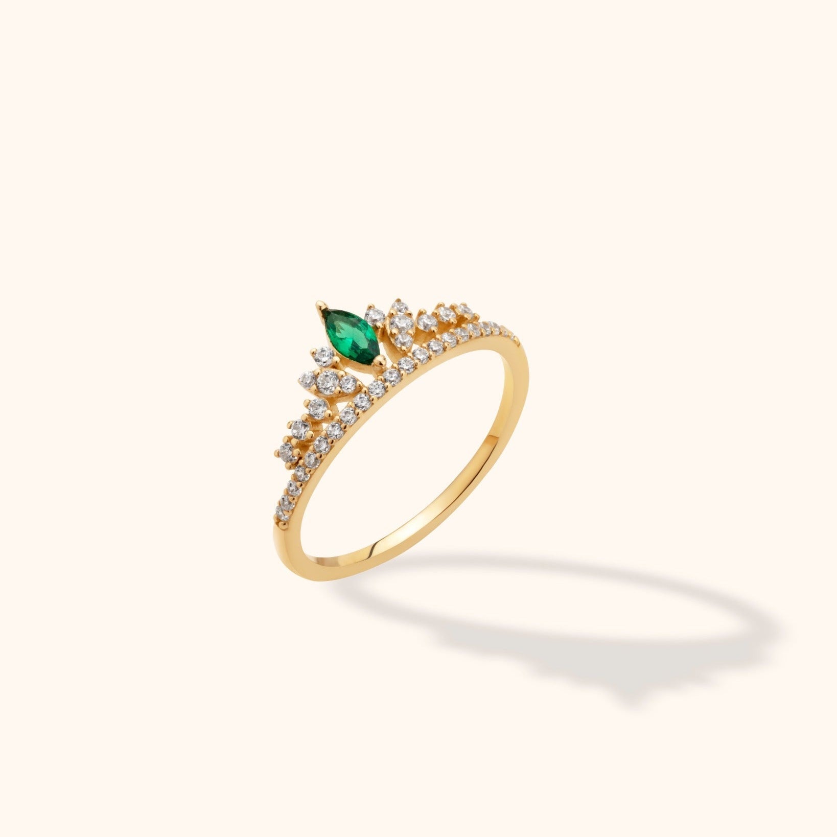Gold-toned tiara-style ring with a marquise-shaped green stone and small clear stones, shown at an angle on a cream background.