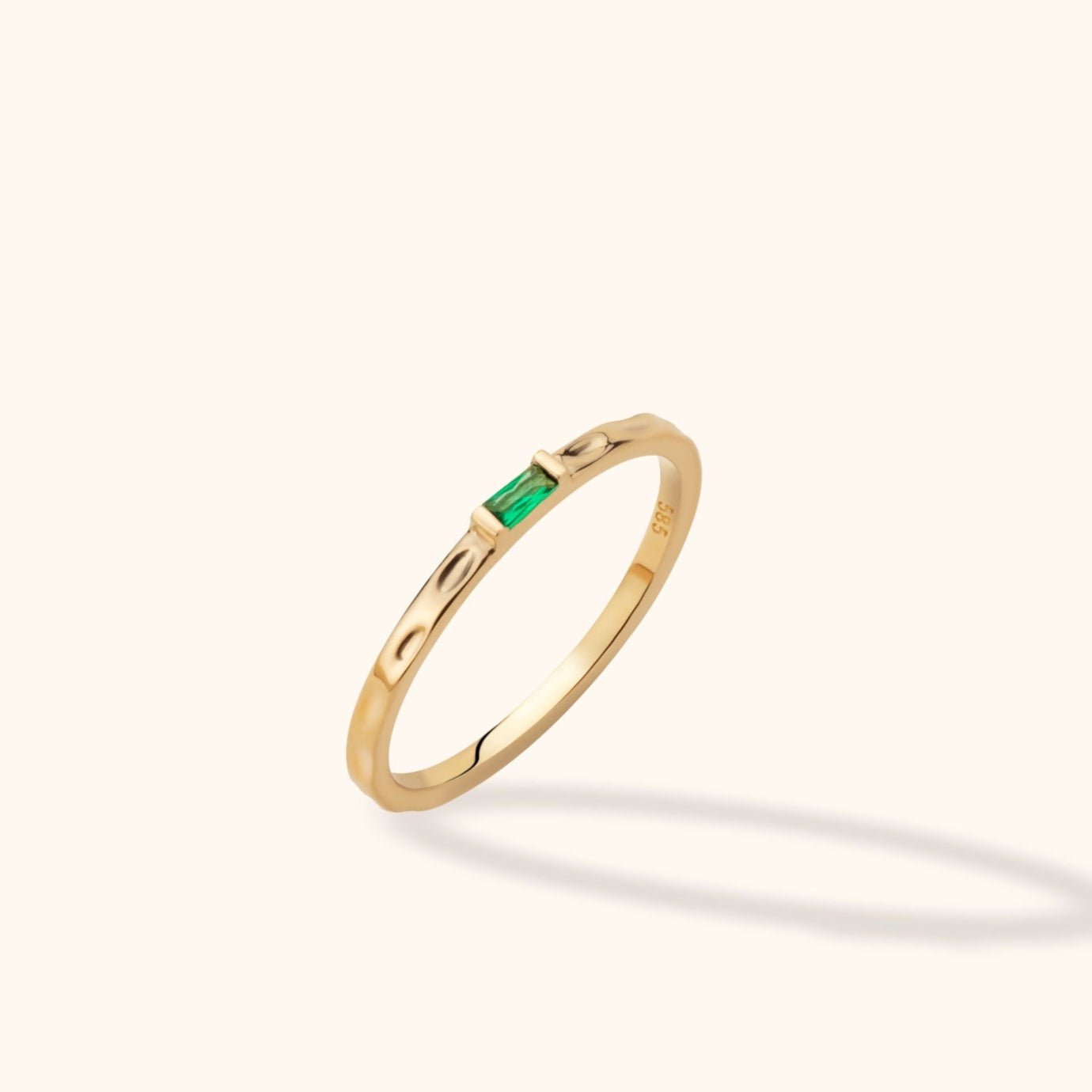Slim gold-toned ring with a rectangular green stone and dimpled band, shown tilted against a pale cream background.
