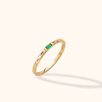Slim gold-toned ring with a rectangular green stone and dimpled band, shown tilted against a pale cream background.
