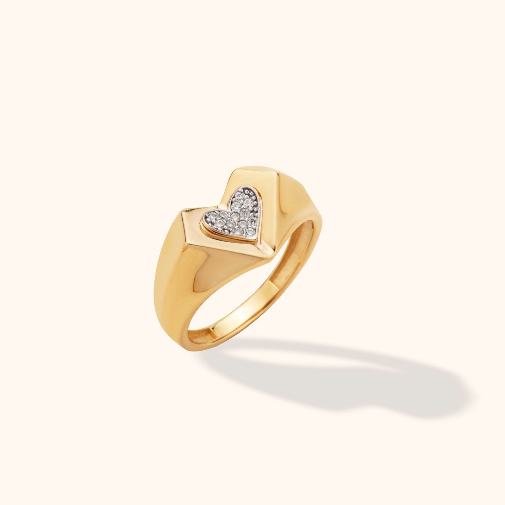 Polished gold-tone signet ring with a broad, tapered band and a heart filled with small clear stones, shown at an angle.