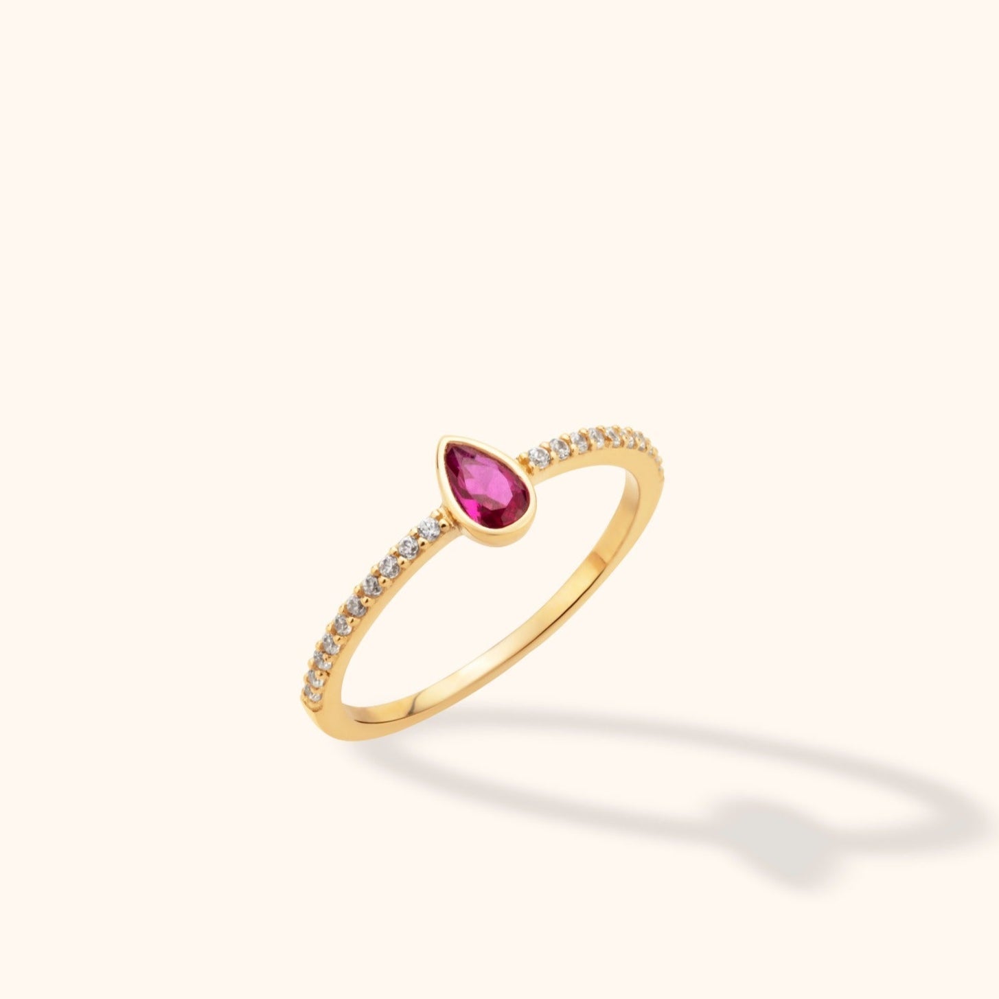 Slim gold-tone ring with a pear-shaped pink center stone and small clear stones along the band, shown at an angle.