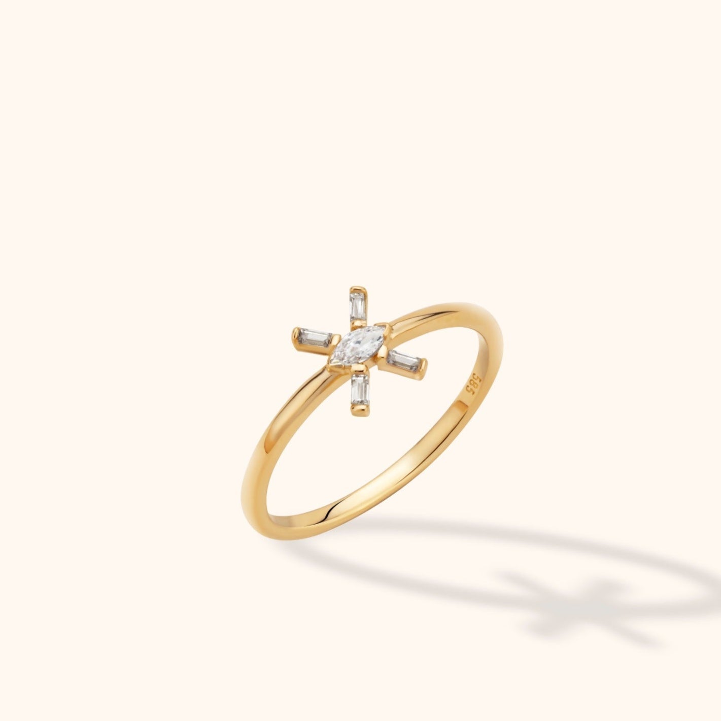 Slim gold-toned ring with a clear marquise-shaped center and four rectangular accents arranged in a starburst on a cream background.