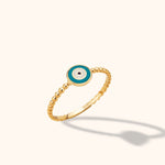 Gold-colored ring with a beaded band and a round turquoise-blue and white eye motif, shown tilted against a pale background.