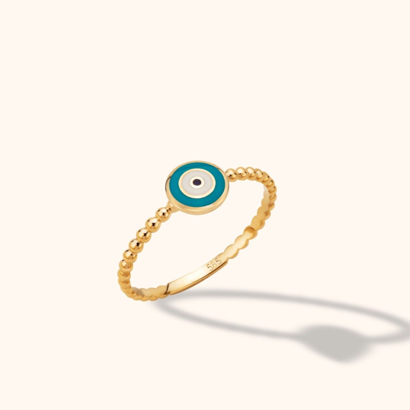 Gold-colored ring with a beaded band and a round turquoise-blue and white eye motif, shown tilted against a pale background.