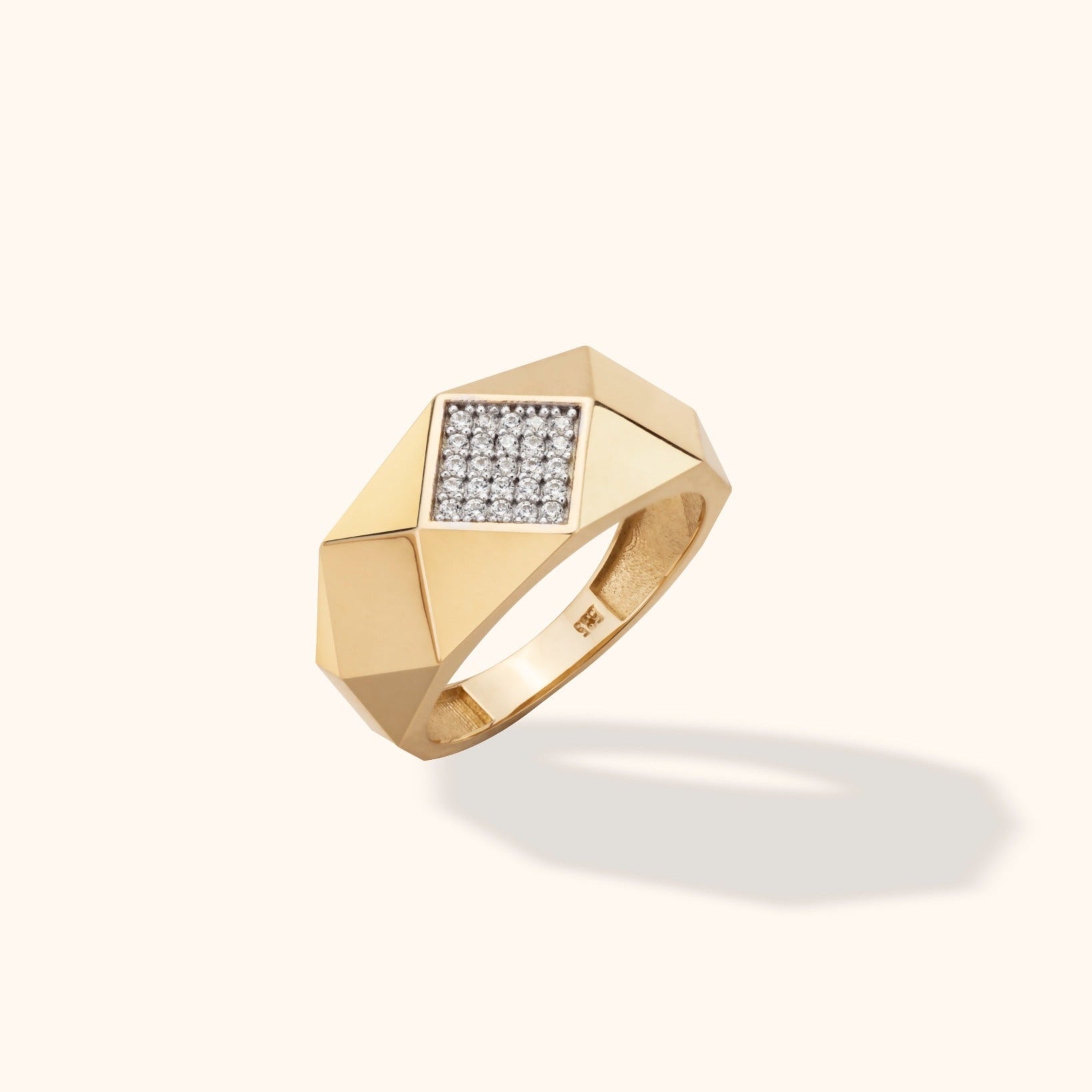 Gold-toned ring with a broad, angular band and a square panel of small clear stones, shown at an angle on a cream background.