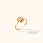Gold-toned ring with a slim band and a ridged seashell-shaped top holding a round white pearl-like bead, on a cream background.