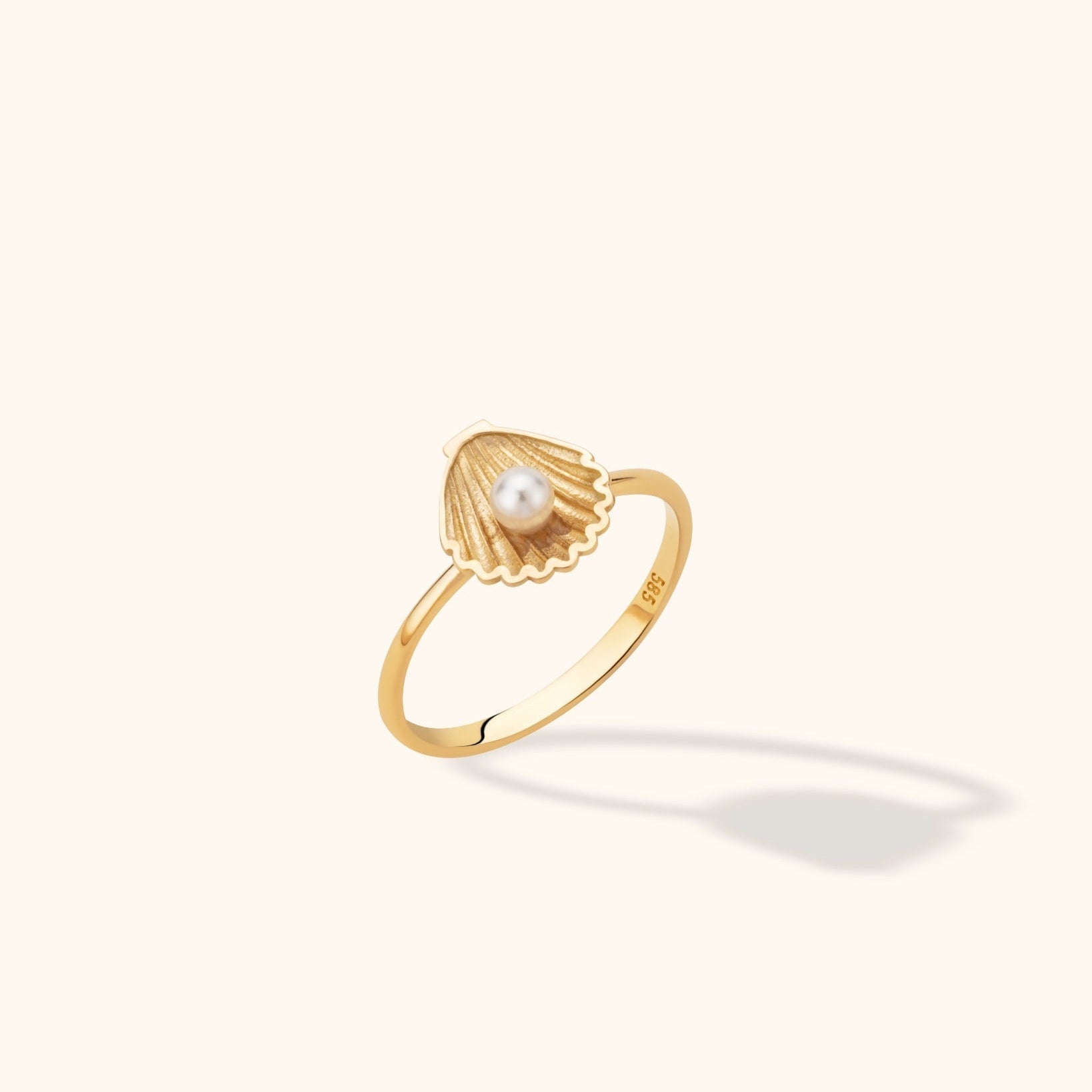 Gold-toned ring with a slim band and a ridged seashell-shaped top holding a round white pearl-like bead, on a cream background.