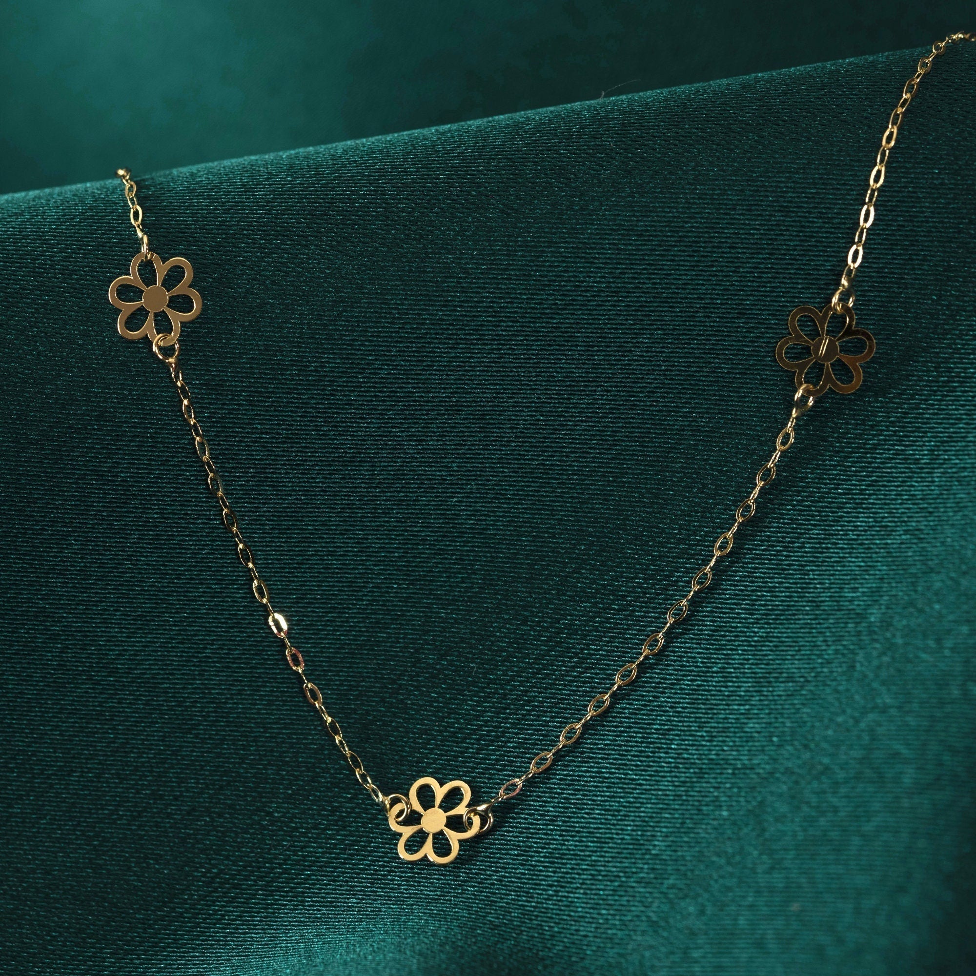 Gold-tone necklace with a fine oval-link chain and three openwork flower motifs, draped over dark green fabric.