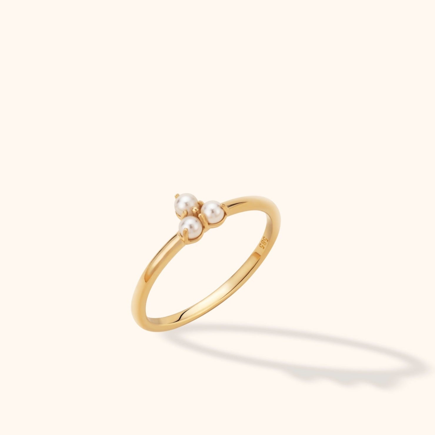 Slim gold-tone ring with three white pearl-like beads in a triangular cluster, shown at an angle on a cream background.