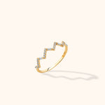 Gold-colored ring with a stepped zigzag front set with round and rectangular clear stones, shown at an angle on a cream background.