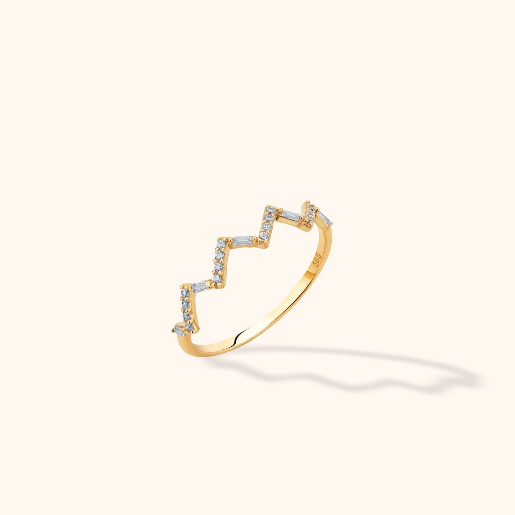 Gold-colored ring with a stepped zigzag front set with round and rectangular clear stones, shown at an angle on a cream background.