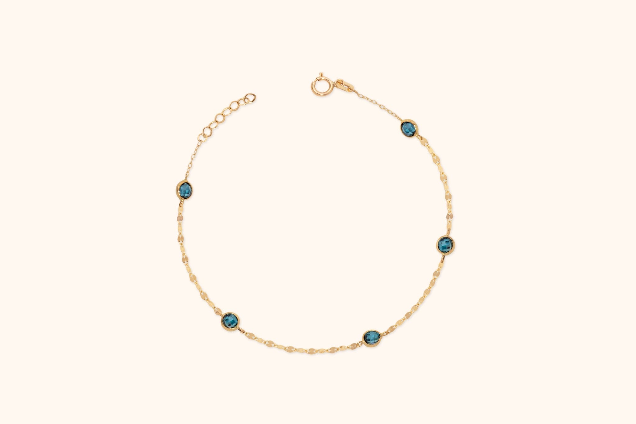 Gold-tone chain bracelet with five round blue faceted accents, a spring-ring clasp and extension links on a cream background.