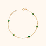 Gold-colored chain bracelet with five round, faceted green accents, a circular clasp and extension links on a cream background.