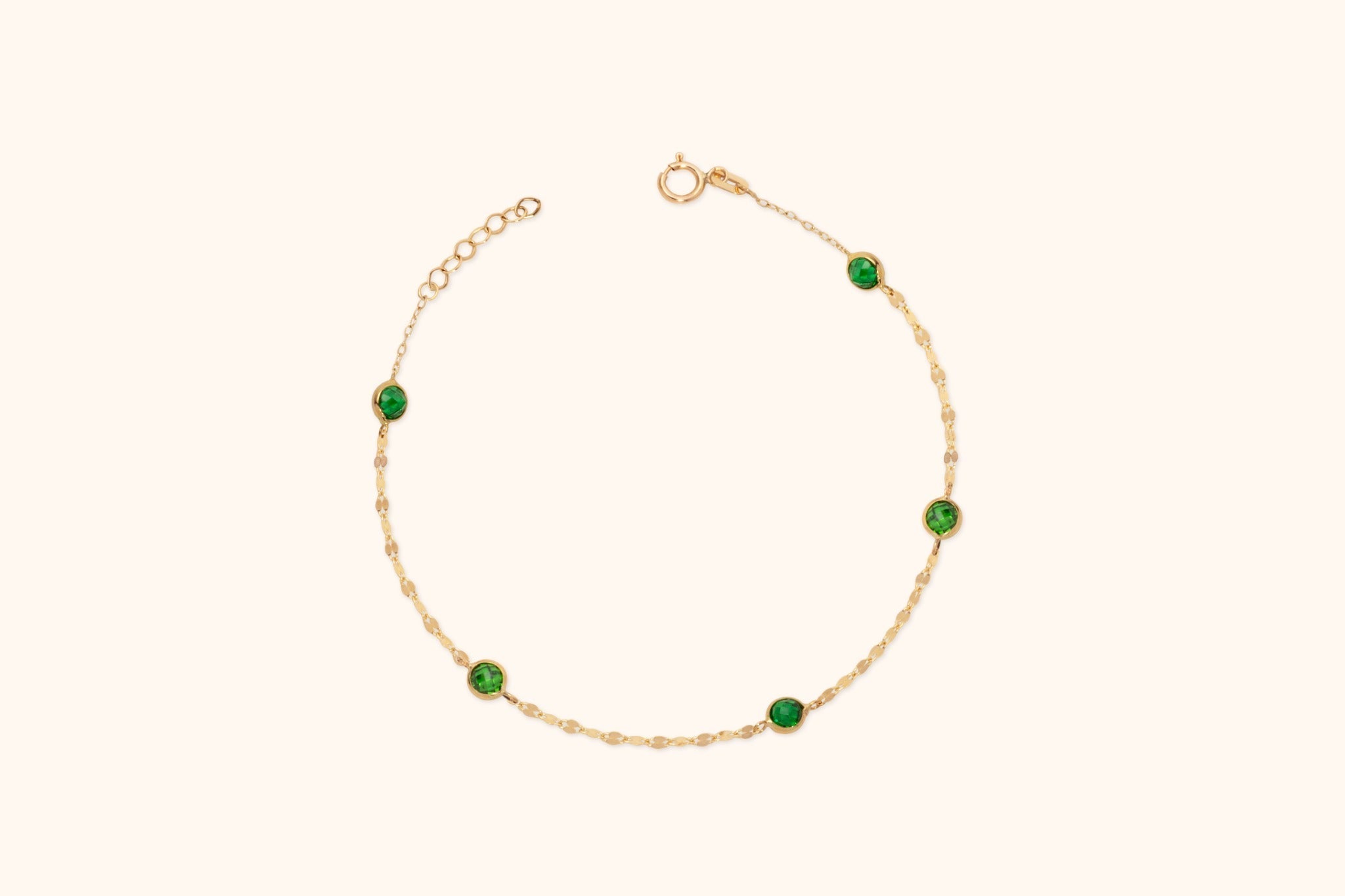 Gold-colored chain bracelet with five round, faceted green accents, a circular clasp and extension links on a cream background.