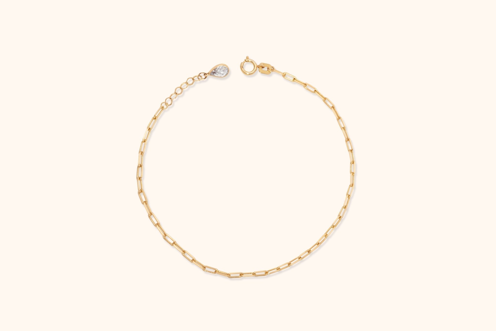 Gold-toned paperclip link bracelet with a round clasp, extension chain and clear teardrop-shaped accent on a cream background.