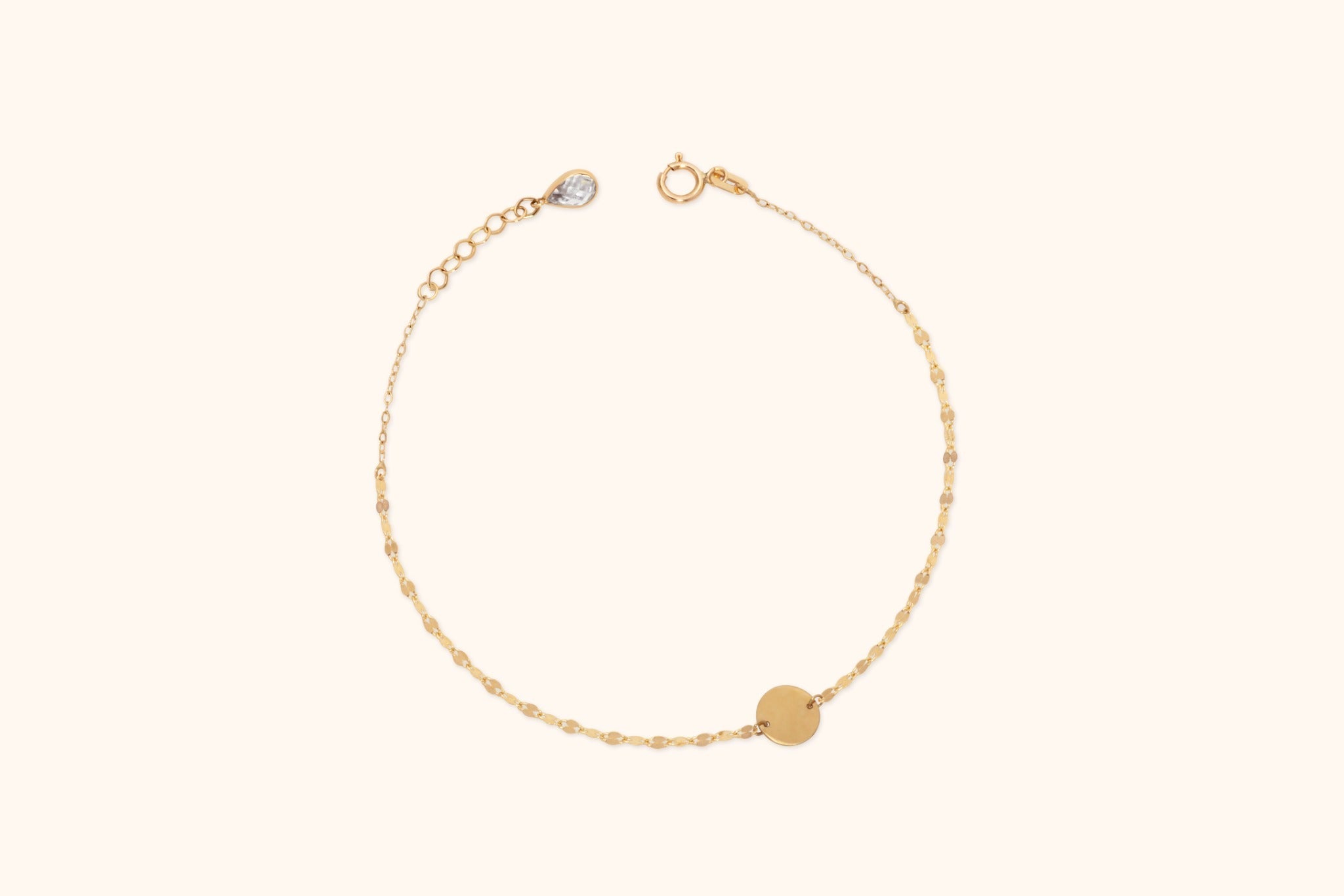 Gold-colored chain bracelet with a flat round disc, spring-ring clasp and extension links, arranged in a circle on a cream background.