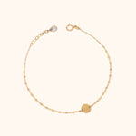 Gold-colored chain bracelet with a flat round disc, spring-ring clasp and extension links, arranged in a circle on a cream background.