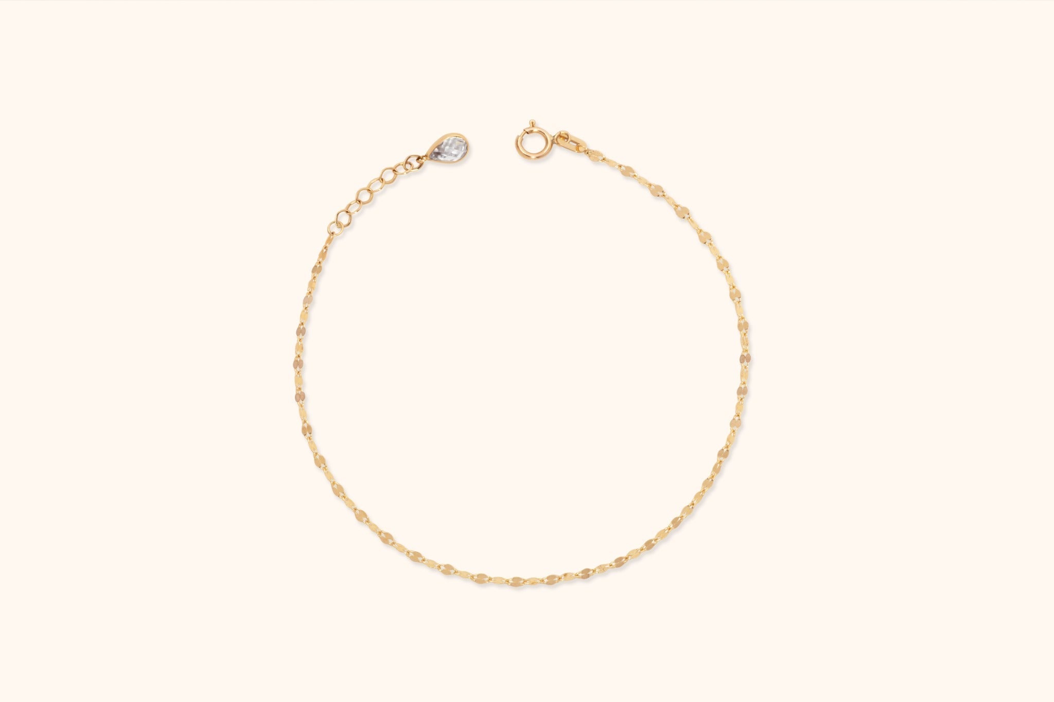 Fine gold-tone chain bracelet with flat oval links, a round clasp, extension chain and clear teardrop-shaped charm on a cream background.