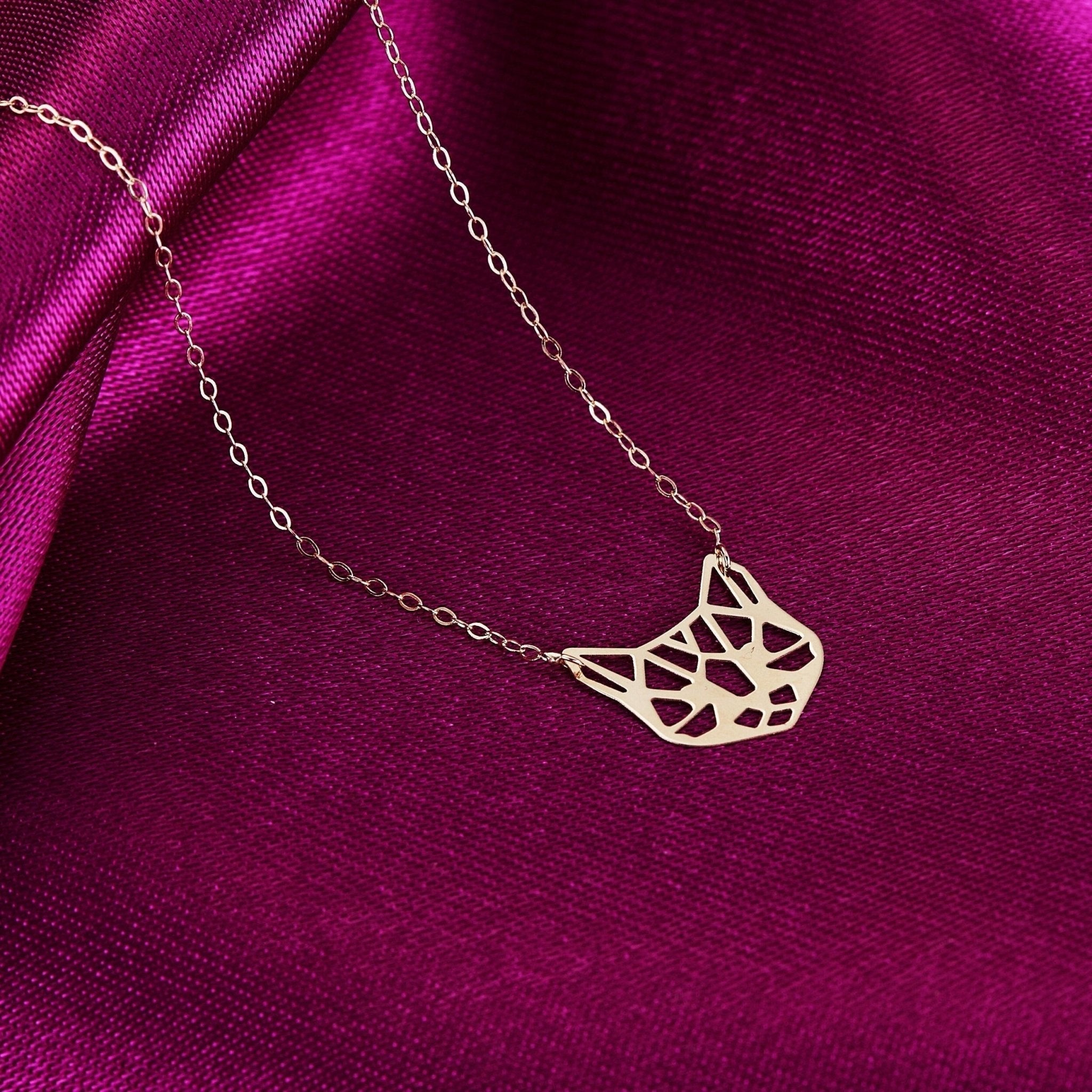 Gold-tone necklace with an openwork geometric cat face pendant and oval-link chain on folded magenta fabric.