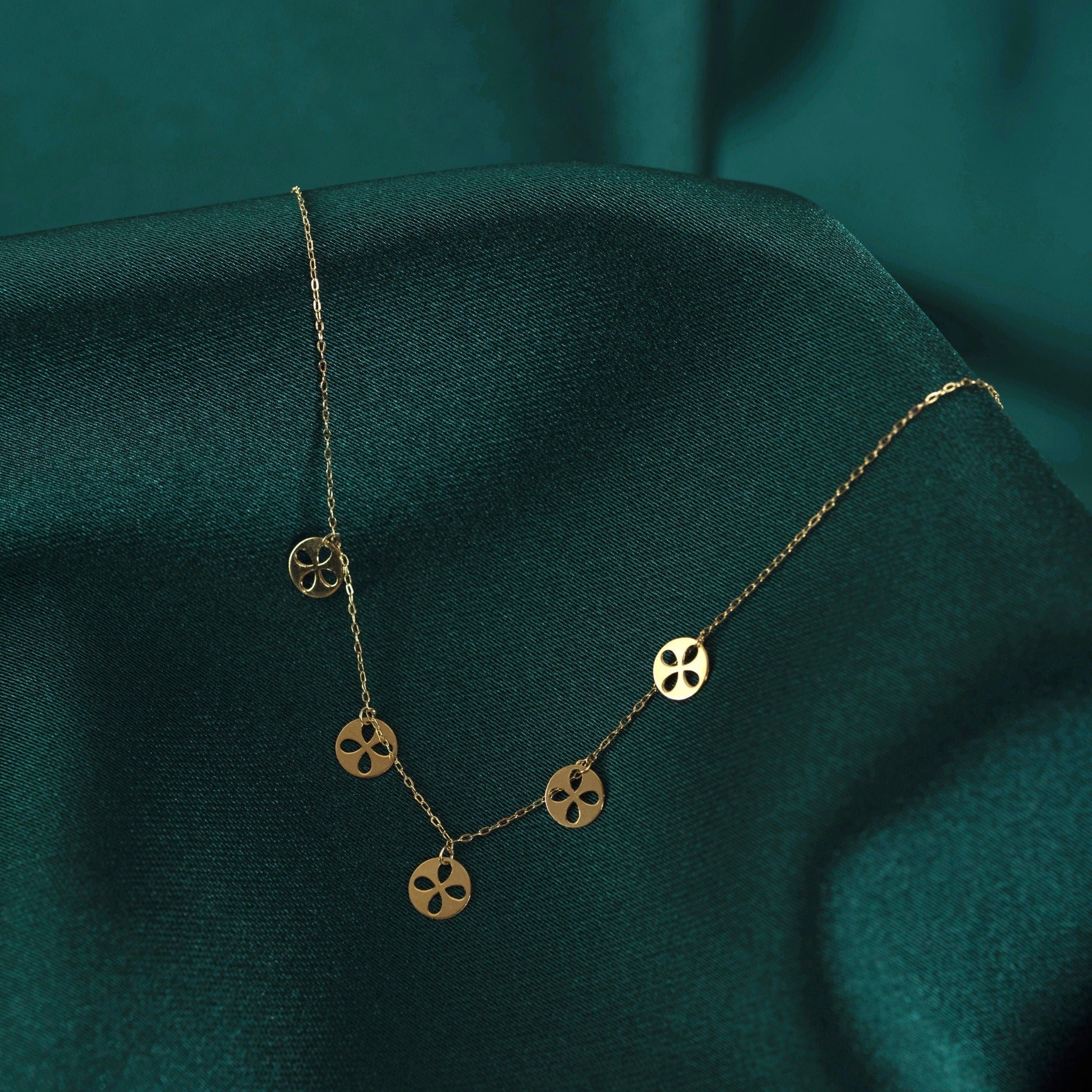 Gold-tone fine chain necklace with five round discs featuring four-petal cutouts, draped over dark green fabric.