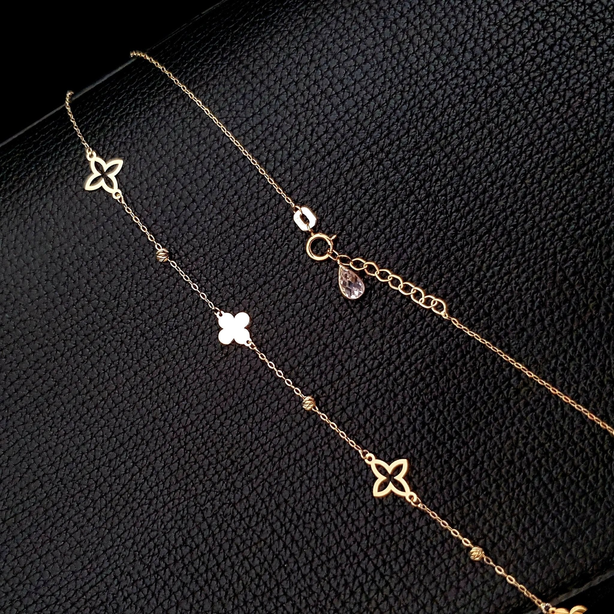 Gold-colored chain necklace with open and solid clover motifs, small textured beads and a round clasp on a black textured surface.